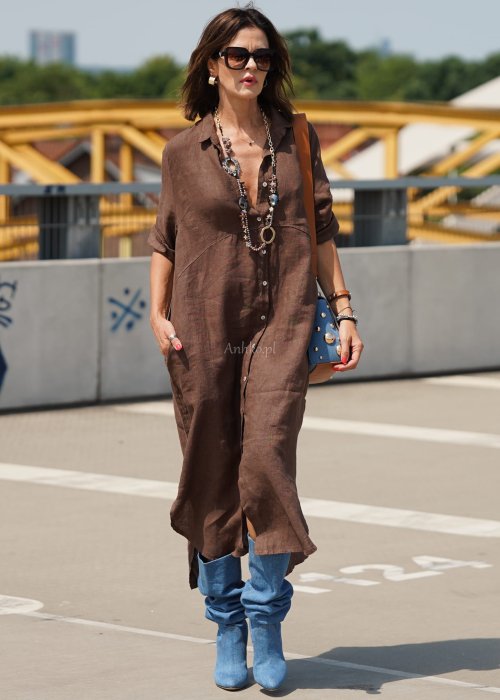 Brązowa lniana sukienka maxi w stylu szmizjerki, zapinana na guziki, o luźnym fasonie oversize, stylizowana z wysokimi kozakami i torebką.