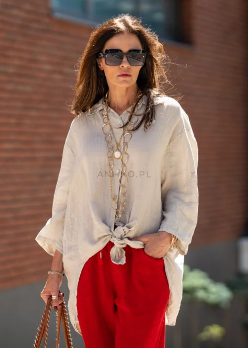 Beżowa lniana koszula damska oversize z asymetrycznym dołem i podpinanymi rękawami, zestawiona z czerwonymi spodniami w eleganckiej stylizacji casual.