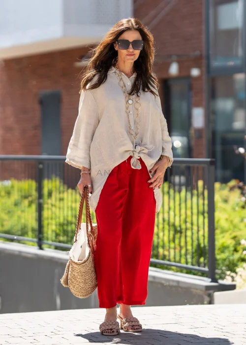 Beżowa lniana koszula damska oversize z asymetrycznym dołem i podpinanymi rękawami, zestawiona z czerwonymi spodniami w eleganckiej stylizacji casual.
