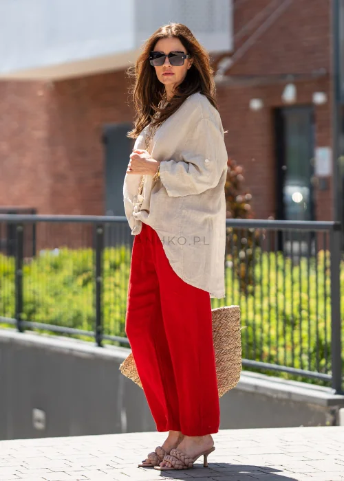 Beżowa lniana koszula damska oversize z asymetrycznym dołem i podpinanymi rękawami, zestawiona z czerwonymi spodniami w eleganckiej stylizacji casual.
