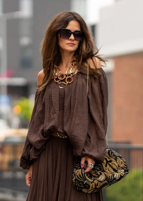 Brązowa bluzka hiszpanka oversize z gniecionej bawełny i szerokim dekoltem odsłaniającym ramiona. Stylizacja boho z długą spódnicą, dużą biżuterią i eleganckimi dodatkami.