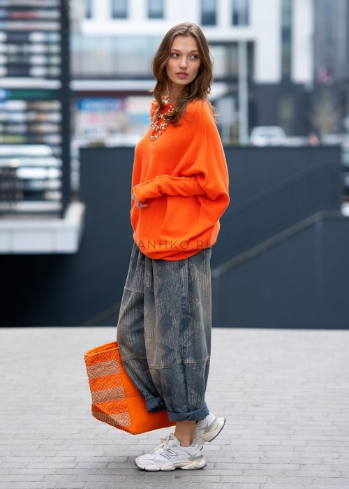 Pomarańczowy sweter damski oversize o nietoperzowym kroju z asymetrycznym dołem, stylizacja miejska z biżuterią i spodniami.