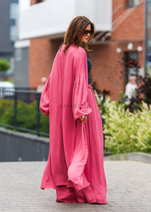 Różowe kimono damskie maxi z długim, płynnie układającym się fasonem i bufiastymi rękawami. Model pokazany w stylizacji z topem i szerokimi spodniami, w nowoczesnym miejskim wydaniu.