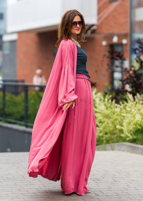 Różowe kimono damskie maxi z długim, płynnie układającym się fasonem i bufiastymi rękawami. Model pokazany w stylizacji z topem i szerokimi spodniami, w nowoczesnym miejskim wydaniu.