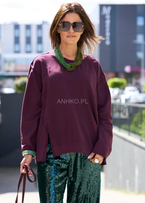 Śliwkowa bluza damska oversize z długim rękawem i okrągłym dekoltem, zestawiona z zieloną spódnicą i biżuterią w stylu miejskim.