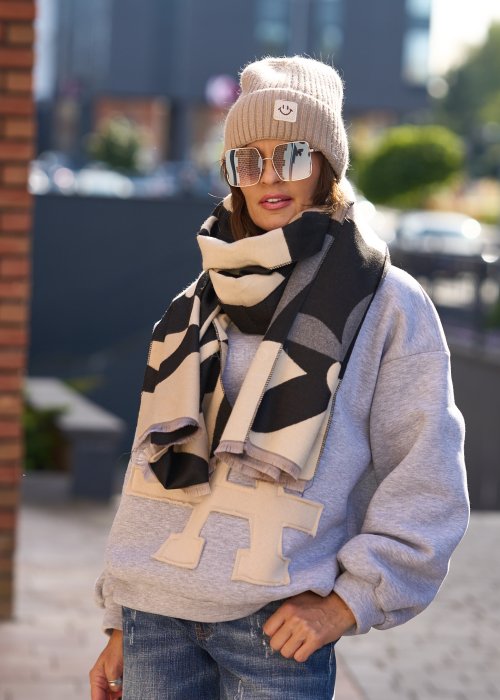 Stylizacja jesienna: beżowa czapka beanie z naszywką uśmiechu oraz duży szalik w beżowo-czarny wzór graficzny, połączone z szarą bluzą oversize i jeansami