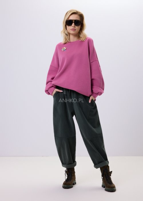 Różowa bluza damska oversize z ozdobną broszką przy dekolcie, o luźnym kroju i obniżonej linii ramion, zestawiona z ciemnymi spodniami w miejskiej stylizacji.