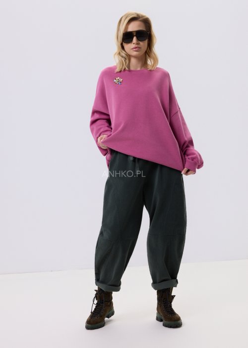 Różowa bluza damska oversize z ozdobną broszką przy dekolcie, o luźnym kroju i obniżonej linii ramion, zestawiona z ciemnymi spodniami w miejskiej stylizacji.