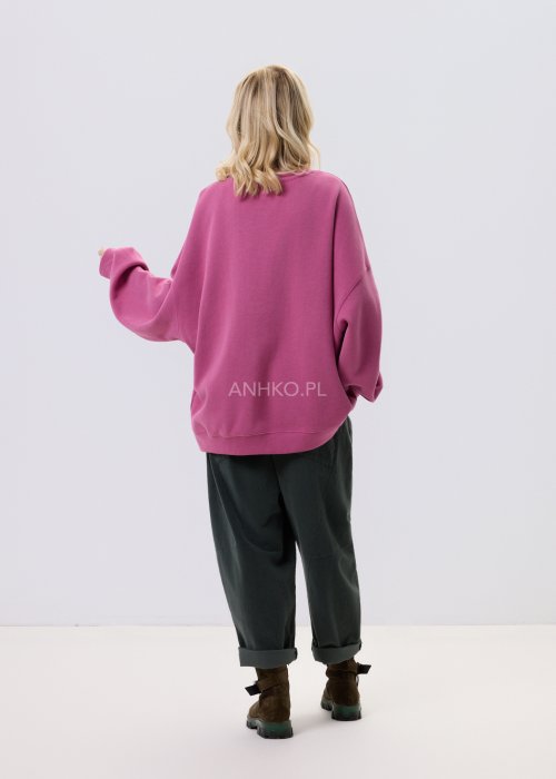 Różowa bluza damska oversize z ozdobną broszką przy dekolcie, o luźnym kroju i obniżonej linii ramion, zestawiona z ciemnymi spodniami w miejskiej stylizacji.