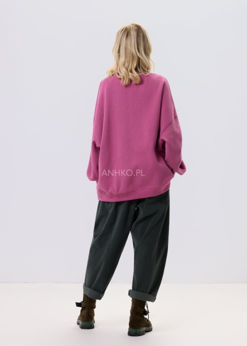 Różowa bluza damska oversize z ozdobną broszką przy dekolcie, o luźnym kroju i obniżonej linii ramion, zestawiona z ciemnymi spodniami w miejskiej stylizacji.