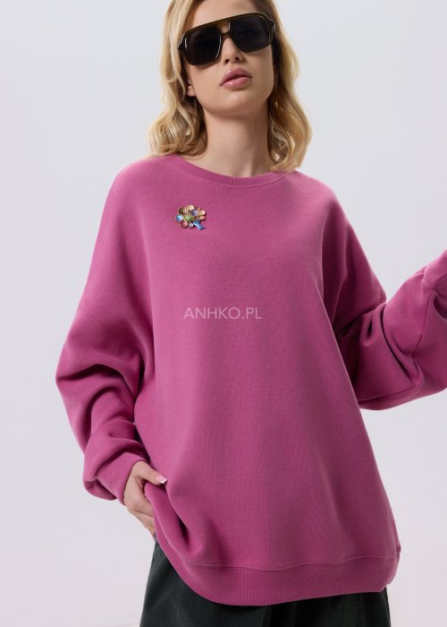 Różowa bluza damska oversize z ozdobną broszką przy dekolcie, o luźnym kroju i obniżonej linii ramion, zestawiona z ciemnymi spodniami w miejskiej stylizacji.