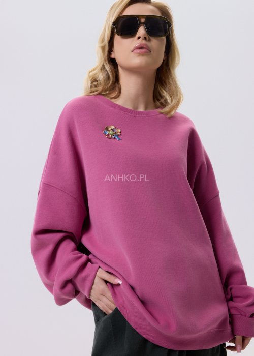 Różowa bluza damska oversize z ozdobną broszką przy dekolcie, o luźnym kroju i obniżonej linii ramion, zestawiona z ciemnymi spodniami w miejskiej stylizacji.
