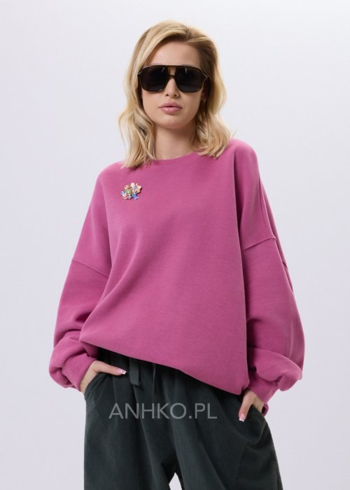 Różowa bluza damska oversize z ozdobną broszką przy dekolcie, o luźnym kroju i obniżonej linii ramion, zestawiona z ciemnymi spodniami w miejskiej stylizacji.