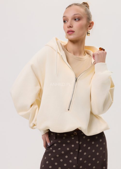 Kremowa damska bluza Creamy Layer o luźnym kroju, z kapturem i krótkim zamkiem przy dekolcie. Model z obniżoną linią ramion i ściągaczami przy rękawach, w stylizacji z brązowymi damskimi spodniami w grochy.
