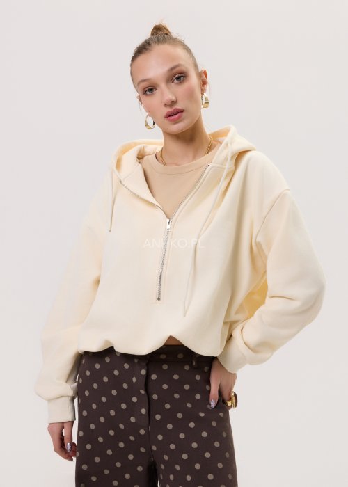 Kremowa damska bluza Creamy Layer o luźnym kroju, z kapturem i krótkim zamkiem przy dekolcie. Model z obniżoną linią ramion i ściągaczami przy rękawach, w stylizacji z brązowymi damskimi spodniami w grochy.