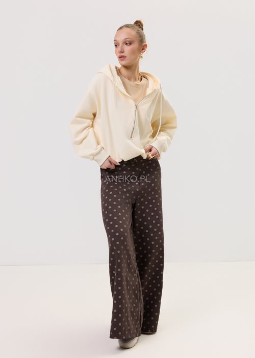 Kremowa damska bluza Creamy Layer o luźnym kroju, z kapturem i krótkim zamkiem przy dekolcie. Model z obniżoną linią ramion i ściągaczami przy rękawach, w stylizacji z brązowymi damskimi spodniami w grochy.