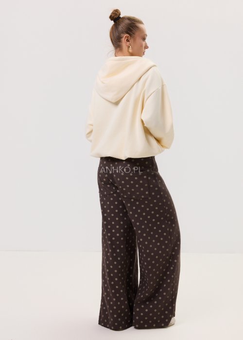 Kremowa damska bluza Creamy Layer o luźnym kroju, z kapturem i krótkim zamkiem przy dekolcie. Model z obniżoną linią ramion i ściągaczami przy rękawach, w stylizacji z brązowymi damskimi spodniami w grochy.