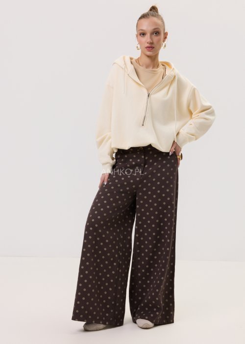 Kremowa damska bluza Creamy Layer o luźnym kroju, z kapturem i krótkim zamkiem przy dekolcie. Model z obniżoną linią ramion i ściągaczami przy rękawach, w stylizacji z brązowymi damskimi spodniami w grochy.