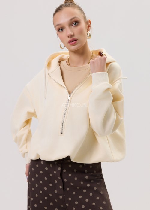 Kremowa damska bluza Creamy Layer o luźnym kroju, z kapturem i krótkim zamkiem przy dekolcie. Model z obniżoną linią ramion i ściągaczami przy rękawach, w stylizacji z brązowymi damskimi spodniami w grochy.
