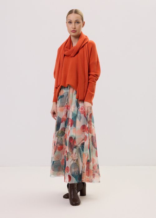 Zwiewna spódnica midi Lunareth Cherry z transparentnej tkaniny w print z wiśniami i liśćmi, z elastyczną gumką w pasie. Stylizacja z pomarańczowym swetrem i brązowymi botkami na obcasie.