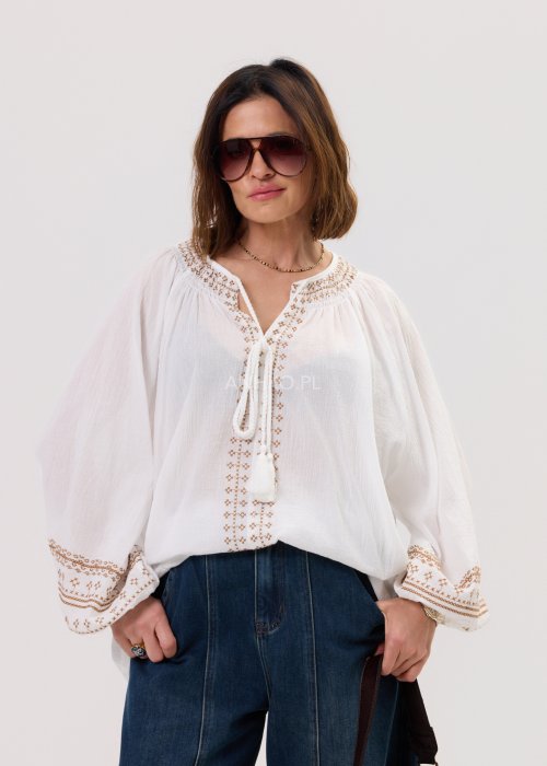 Damska bluzka Boho Rosa biała z delikatnym haftem przy dekolcie i mankietach, wiązaniem z chwostami oraz luźnym, zwiewnym fasonem w stylizacji z jeansami.