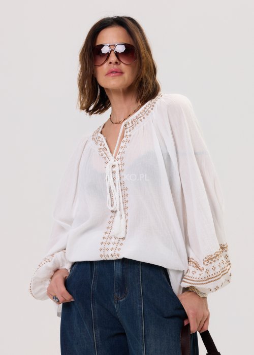 Damska bluzka Boho Rosa biała z delikatnym haftem przy dekolcie i mankietach, wiązaniem z chwostami oraz luźnym, zwiewnym fasonem w stylizacji z jeansami.
