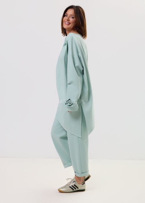 Damskie miętowe spodnie Starola Rose z podwiniętą nogawką i eleganckim paskiem, luźniejszy fason z zakładkami z przodu, w casualowej stylizacji ze sneakersami.