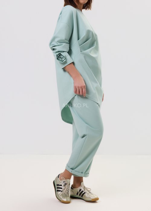 Damskie miętowe spodnie Starola Rose z podwiniętą nogawką i eleganckim paskiem, luźniejszy fason z zakładkami z przodu, w casualowej stylizacji ze sneakersami.