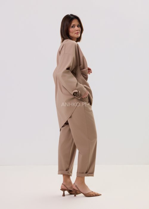 Beżowa bluzka oversize z długim rękawem i asymetrycznym dołem, ozdobiona biżuteryjną broszką w talii, w eleganckiej minimalistycznej stylizacji.