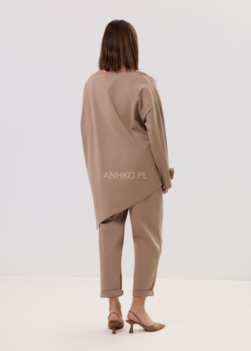 Beżowa bluzka oversize z długim rękawem i asymetrycznym dołem, ozdobiona biżuteryjną broszką w talii, w eleganckiej minimalistycznej stylizacji.