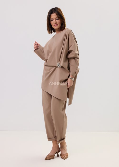 Beżowa bluzka oversize z długim rękawem i asymetrycznym dołem, ozdobiona biżuteryjną broszką w talii, w eleganckiej minimalistycznej stylizacji.