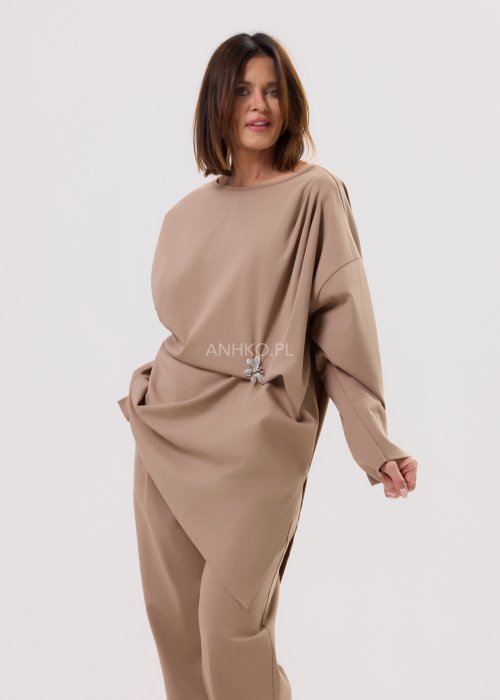 Beżowa bluzka oversize z długim rękawem i asymetrycznym dołem, ozdobiona biżuteryjną broszką w talii, w eleganckiej minimalistycznej stylizacji.
