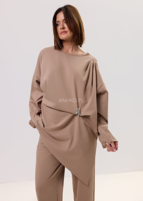 Beżowa bluzka oversize z długim rękawem i asymetrycznym dołem, ozdobiona biżuteryjną broszką w talii, w eleganckiej minimalistycznej stylizacji.