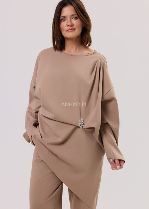 Beżowa bluzka oversize z długim rękawem i asymetrycznym dołem, ozdobiona biżuteryjną broszką w talii, w eleganckiej minimalistycznej stylizacji.
