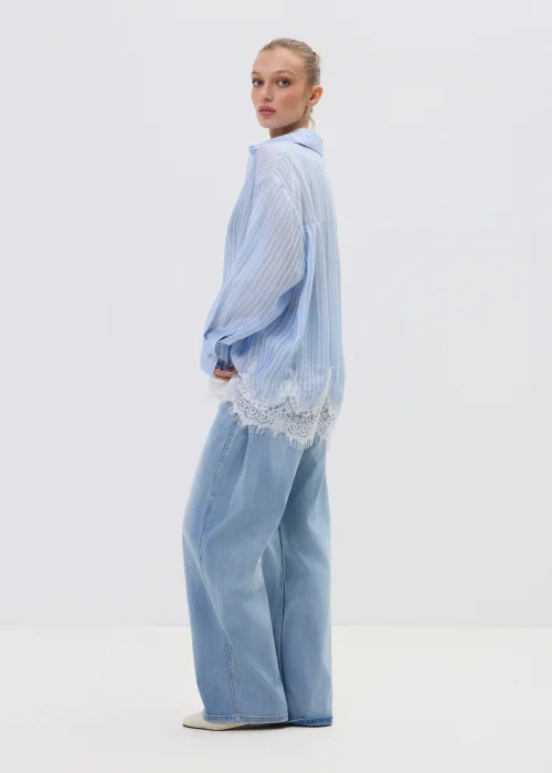 Niebieska koszula damska w delikatne paski z koronkowym wykończeniem u dołu, lekko transparentna, o luźnym kroju w stylizacji casual z jeansami.