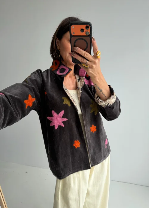 Żakiet boho w grafitowym kolorze ozdobiony kolorowymi haftowanymi aplikacjami w kształcie kwiatów i abstrakcyjnych motywów. Krótki model z szerokimi rękawami i dekoracyjną stójką doskonale podkreśla artystyczny charakter stylizacji.