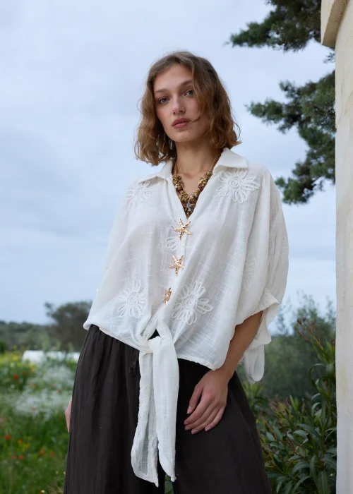 Biała koszula boho z haftowanymi kwiatami i ozdobnymi zapięciami w kształcie rozgwiazd. Zwiewny fason wiązany w talii, zestawiony z ciemną spódnicą maxi.