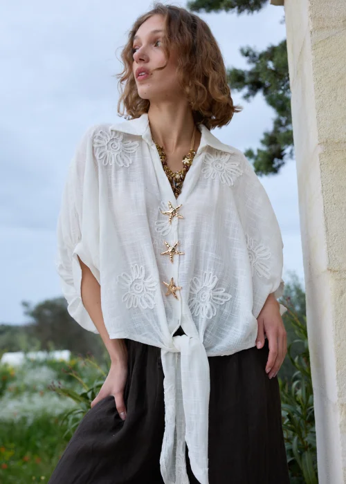 Biała koszula boho z haftowanymi kwiatami i ozdobnymi zapięciami w kształcie rozgwiazd. Zwiewny fason wiązany w talii, zestawiony z ciemną spódnicą maxi.
