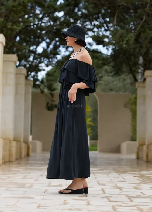 Czarna sukienka maxi hiszpanka z efektownymi falbanami przy dekolcie i regulacją w talii. Elegancka stylizacja z odkrytymi ramionami, kapeluszem i wyrazistą biżuterią w minimalistycznym wydaniu.