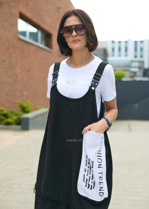 Czarna sukienka ogrodniczka midi z regulowanymi szelkami i kontrastową białą kieszenią z nadrukiem. Luźny model o nowoczesnym, casualowym charakterze, zestawiony z białym T-shirtem i czarnymi botkami.