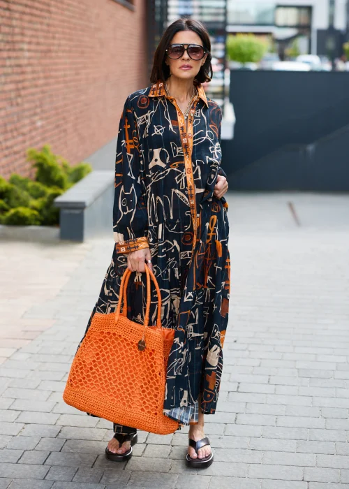 Pomarańczowa ażurowa torebka shopper wykonana z plecionego splotu z krótkimi uchwytami. Lekki, pojemny model doskonale uzupełnia letnią stylizację z czarną sukienką we wzory w odcieniach pomarańczu i beżu.