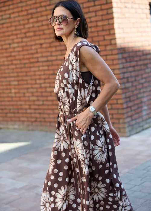 Sukienka maxi Plus Size w brązowym kolorze z dużym kwiatowym wzorem i białymi groszkami, z okrągłym dekoltem, kieszeniami i ozdobnym paskiem w talii, prezentowana w miejskiej stylizacji.