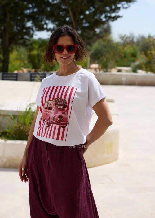 Biały damski T-shirt z czerwonym nadrukiem retro samochodu i pionowymi pasami, w zestawieniu z bordowymi spodniami o luźnym fasonie.