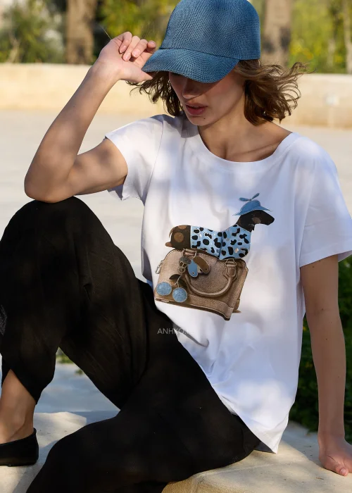 Biały damski T-shirt z kolorowym nadrukiem jamnika w niebieskiej czapce siedzącego na torebce. Luźny fason z krótkim rękawem doskonale komponuje się z czarnymi lnianymi spodniami i codziennymi stylizacjami.