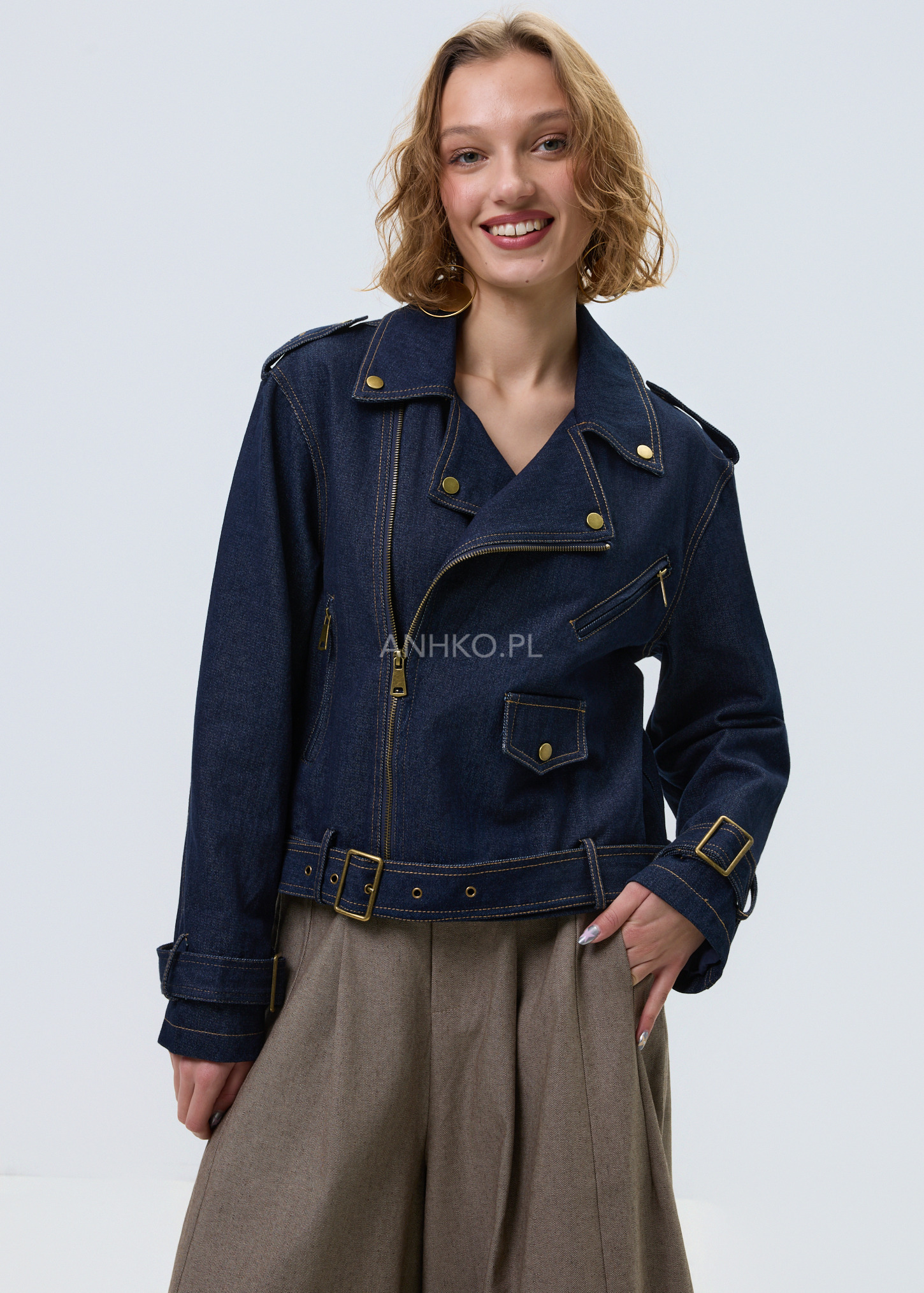 Damska kurtka jeansowa w kolorze granatowego denimu, model typu ramoneska z zamkiem, klapami, ozdobnymi napami i paskiem w pasie, stylizacja casual.